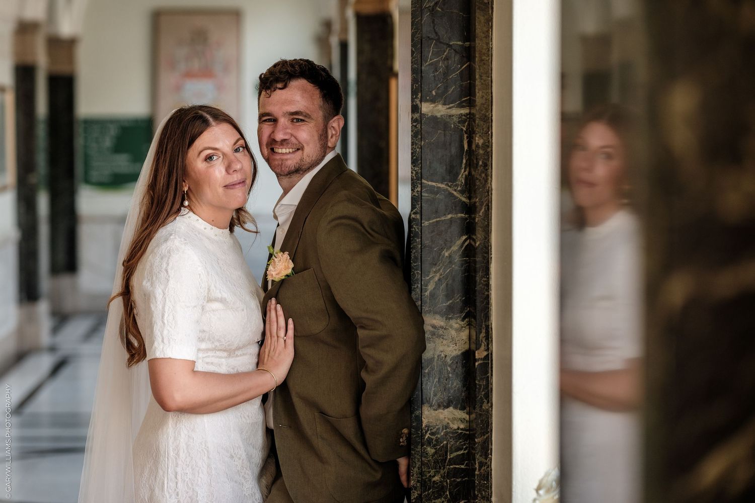 Bride and groom pose elegantly in a marble hallway, the bride in white dress and groom in brown suit with boutonniere.