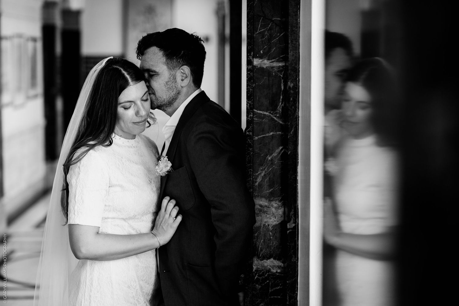 Black and white wedding photo of bride and groom sharing intimate moment, groom kissing bride's head near marble column.