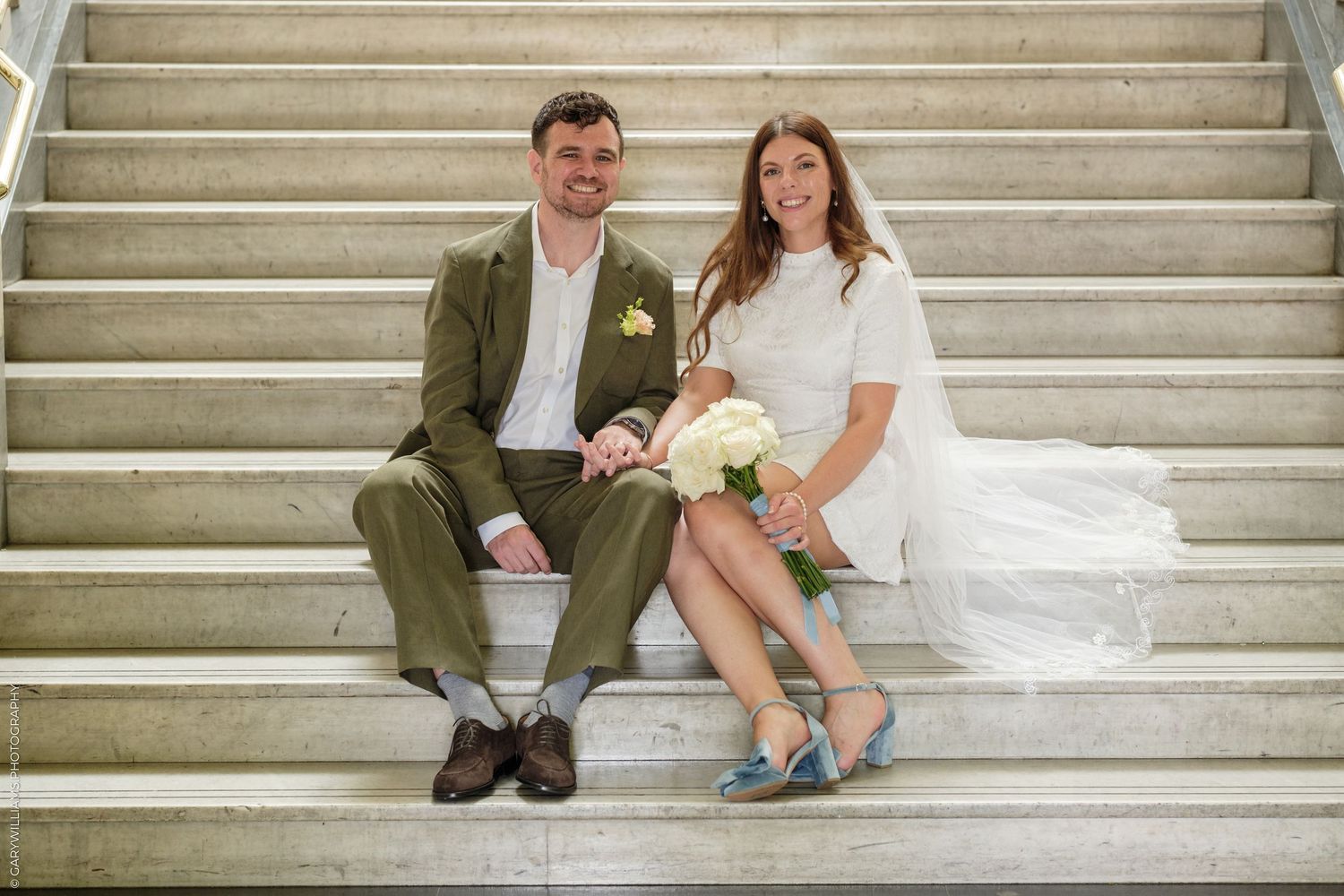 Smiling bride and groom sitting on marble steps, bride holding white bouquet, groom in olive suit, bride in white dress.