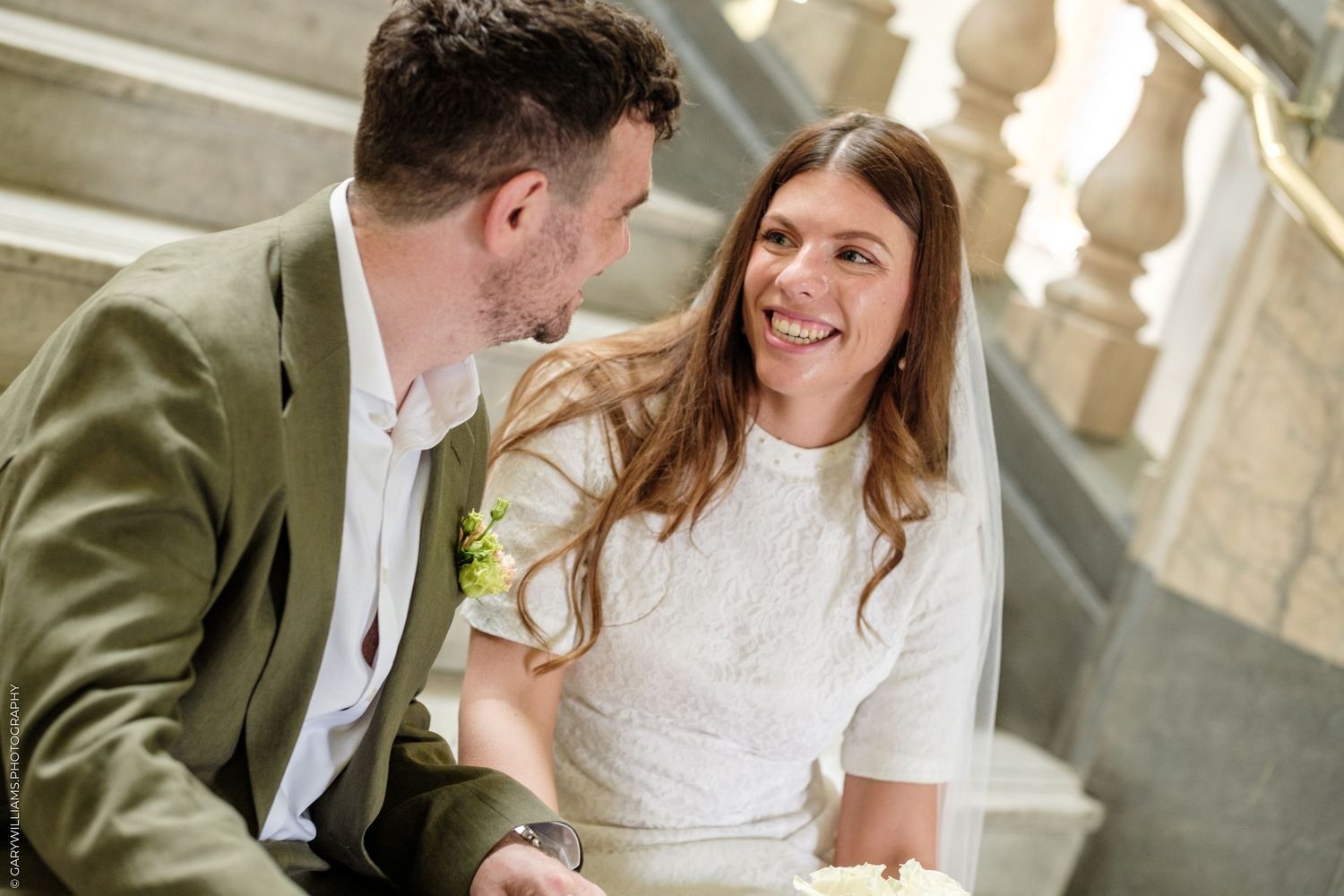 Smiling bride in white dress and veil looks at groom in green suit on elegant staircase.