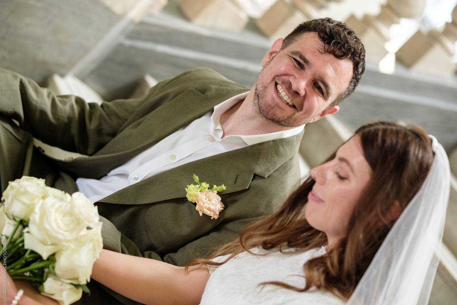Smiling groom in green suit looks at bride holding white bouquet on their wedding day.