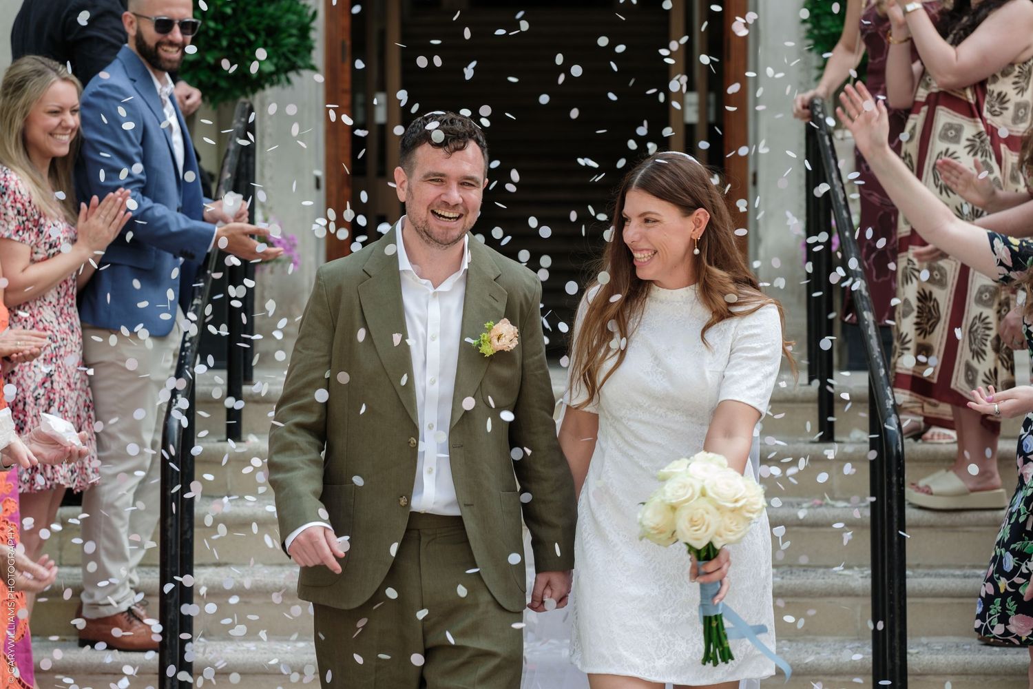 Newlywed couple walking through confetti shower outside wedding venue, bride holding bouquet, groom in olive suit.