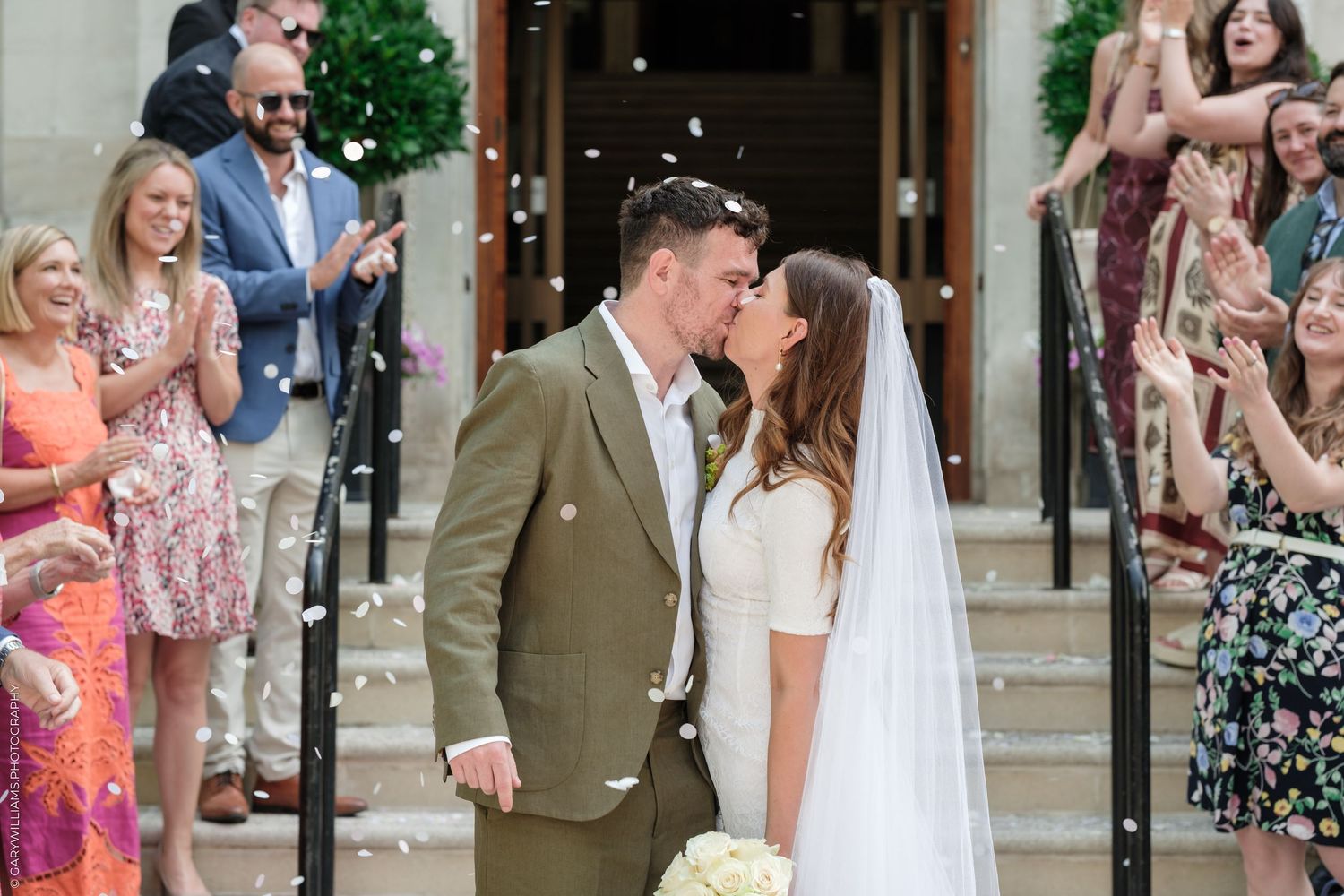 Bride and groom share a kiss on church steps while guests throw white confetti during outdoor wedding celebration.