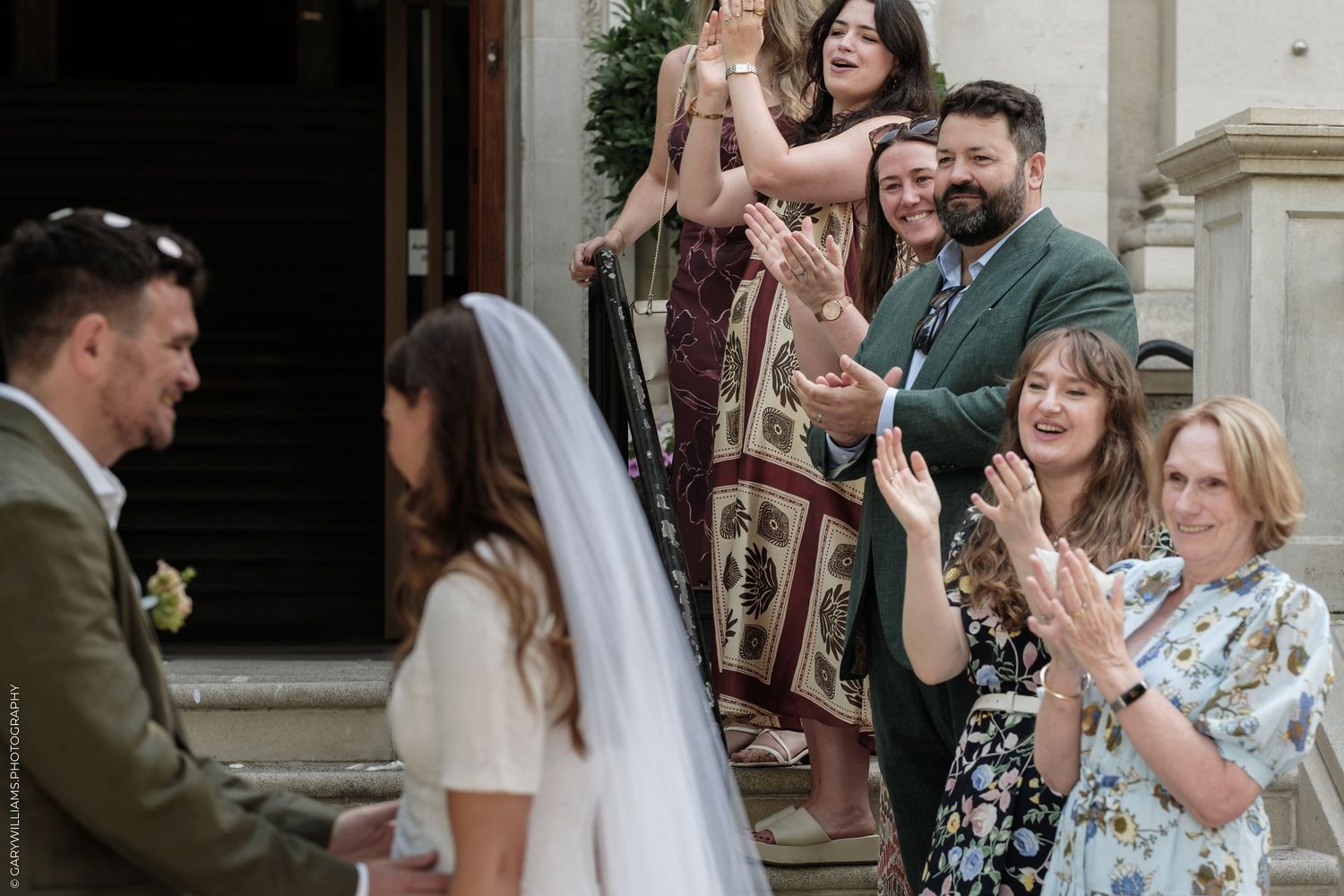 Guests clapping and celebrating as a bride in a white veil walks past on her wedding day outdoors.