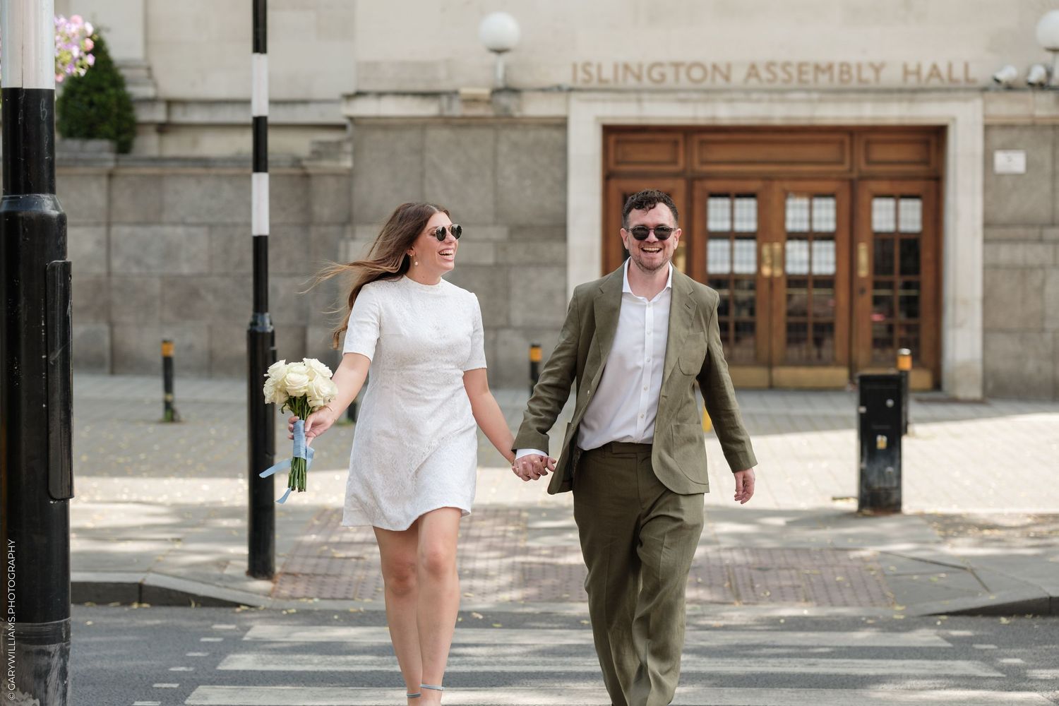 Couple holding hands outside Islington Assembly Hall on their wedding day, bride in white mini dress holding bouquet.