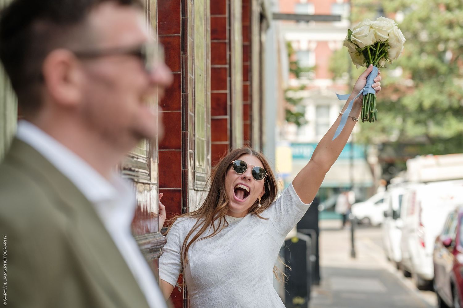 Joyful bride in white dress and sunglasses raising bouquet on city street with groom in foreground.