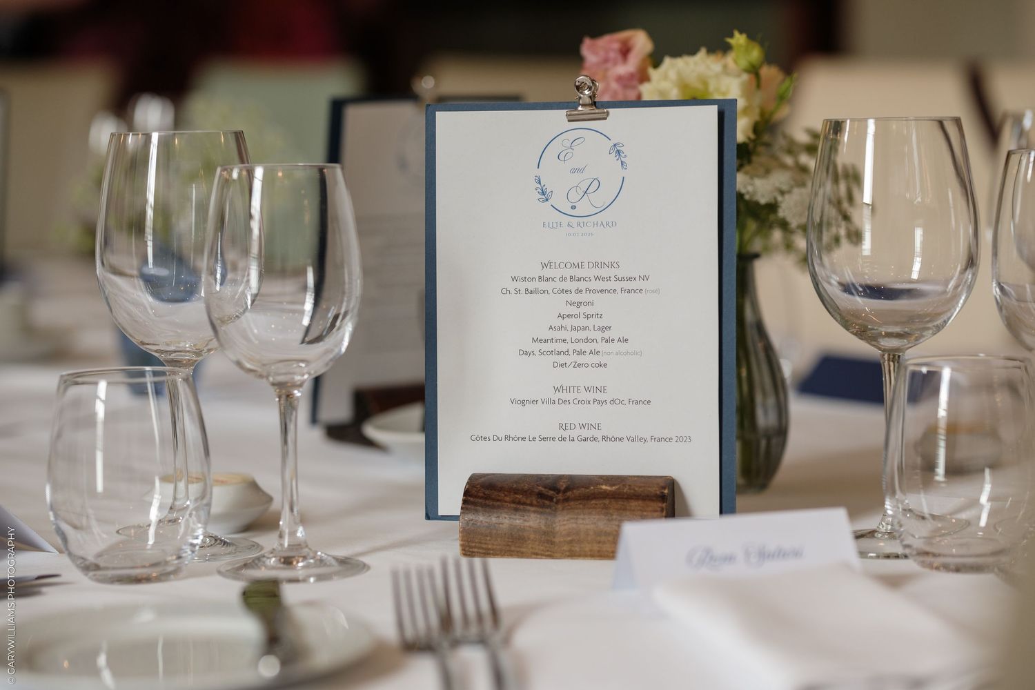 Elegant wedding reception table setting with wine glasses, menu card, and floral centerpiece.