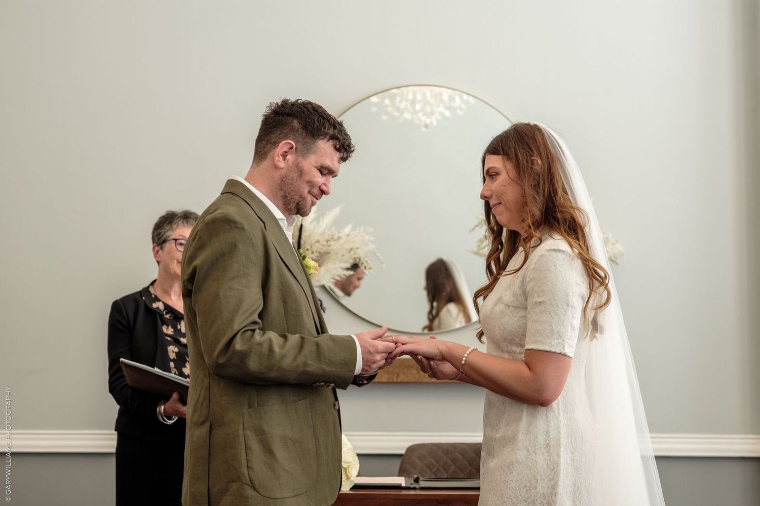 Bride and groom exchange rings during intimate wedding ceremony, officiant standing behind them near arched mirror.