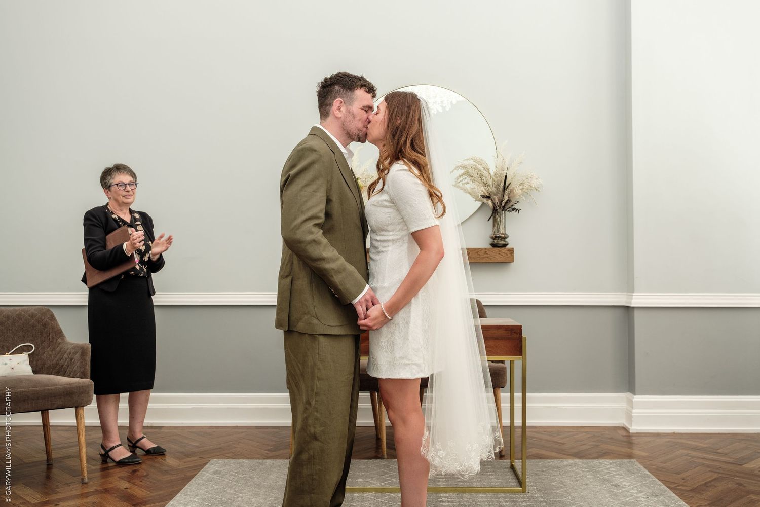 Couple sharing first kiss at wedding ceremony while guest applauds in elegant room with round mirror backdrop.