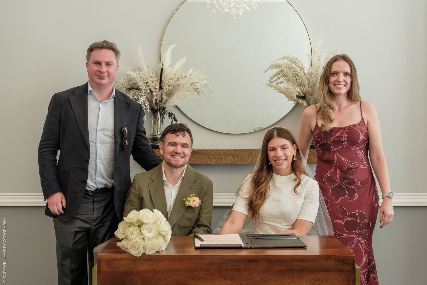 Couple signing wedding register at wooden desk with two witnesses standing behind them in elegant venue.