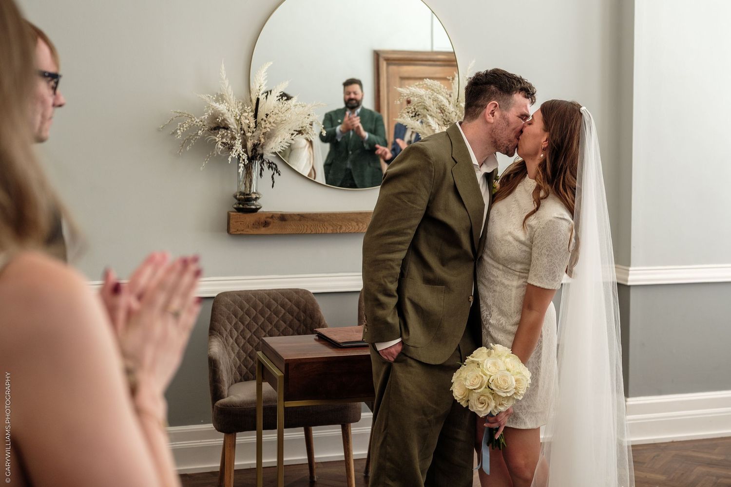 Bride and groom share first kiss at intimate wedding ceremony while guests applaud and photographer captures moment in mirror.