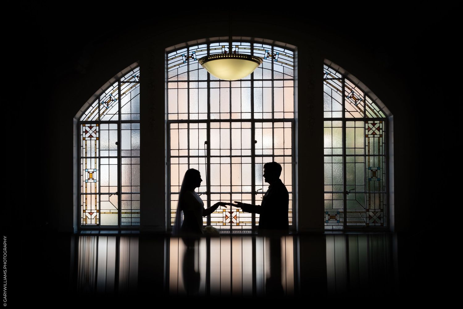 Silhouette of couple exchanging rings before stunning stained glass arched window in elegant venue.