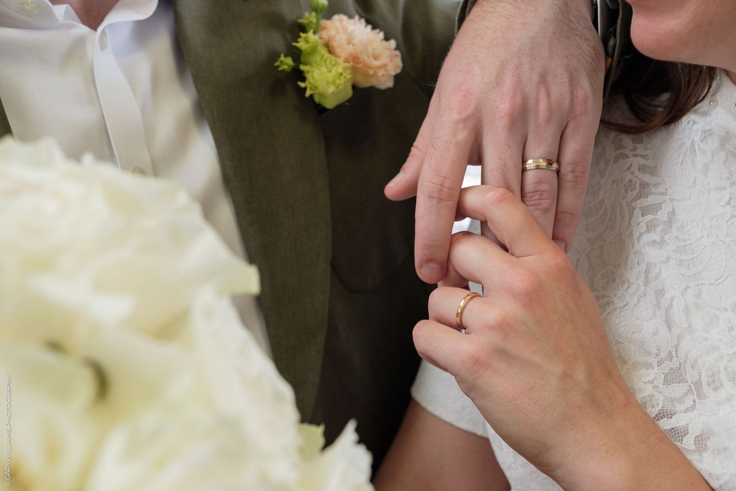 Close-up of a couple exchanging wedding rings during a ceremony, with a white floral bouquet in the foreground.