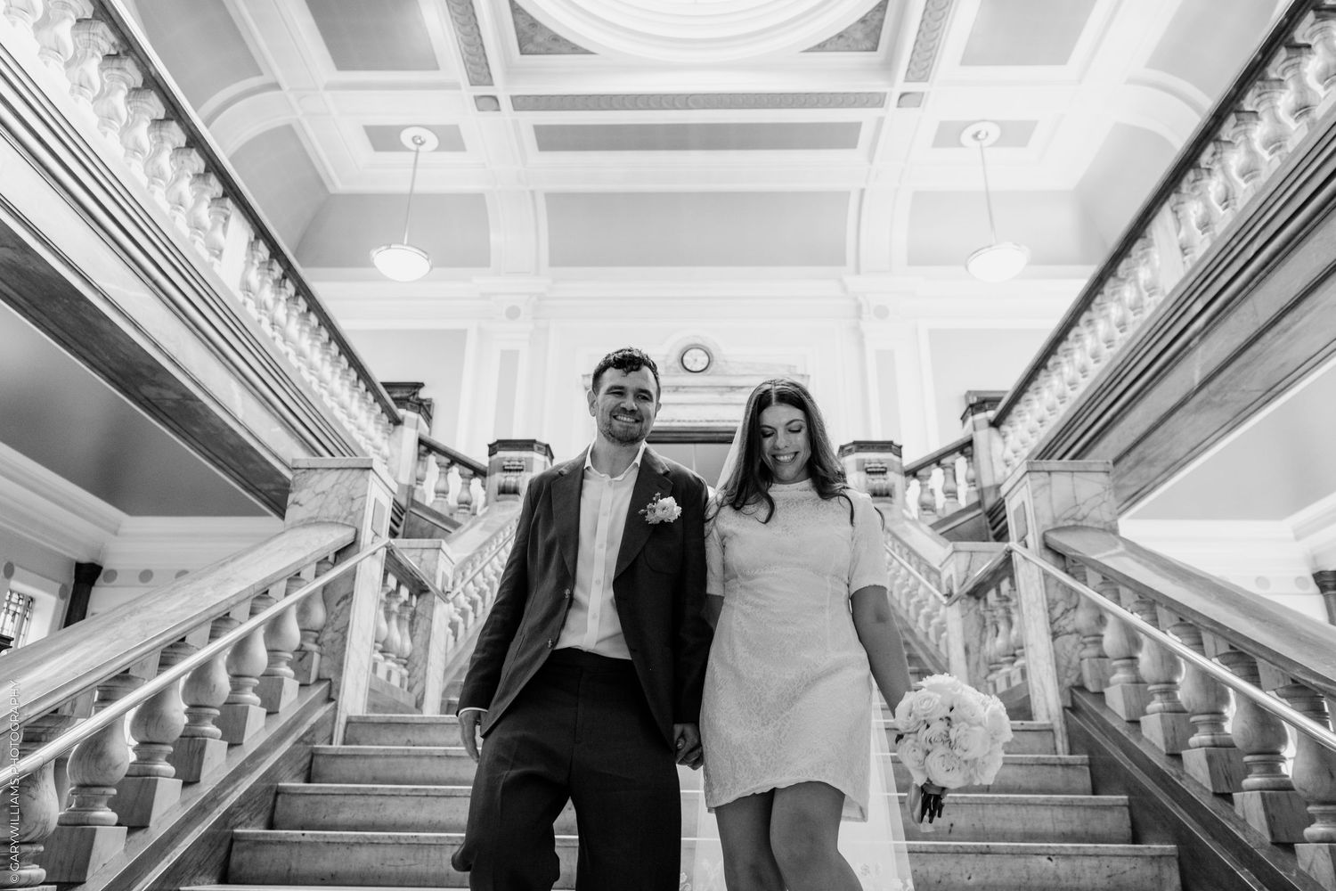 Black and white photo of a bride and groom holding hands on grand staircase in elegant wedding venue.