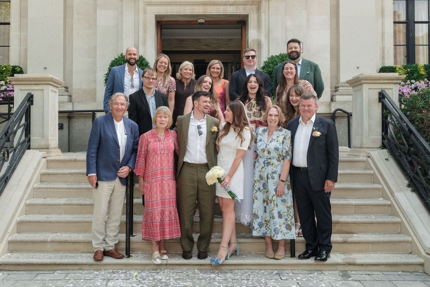 Large family group posing on stone steps outside elegant building, dressed in formal attire for a wedding celebration.