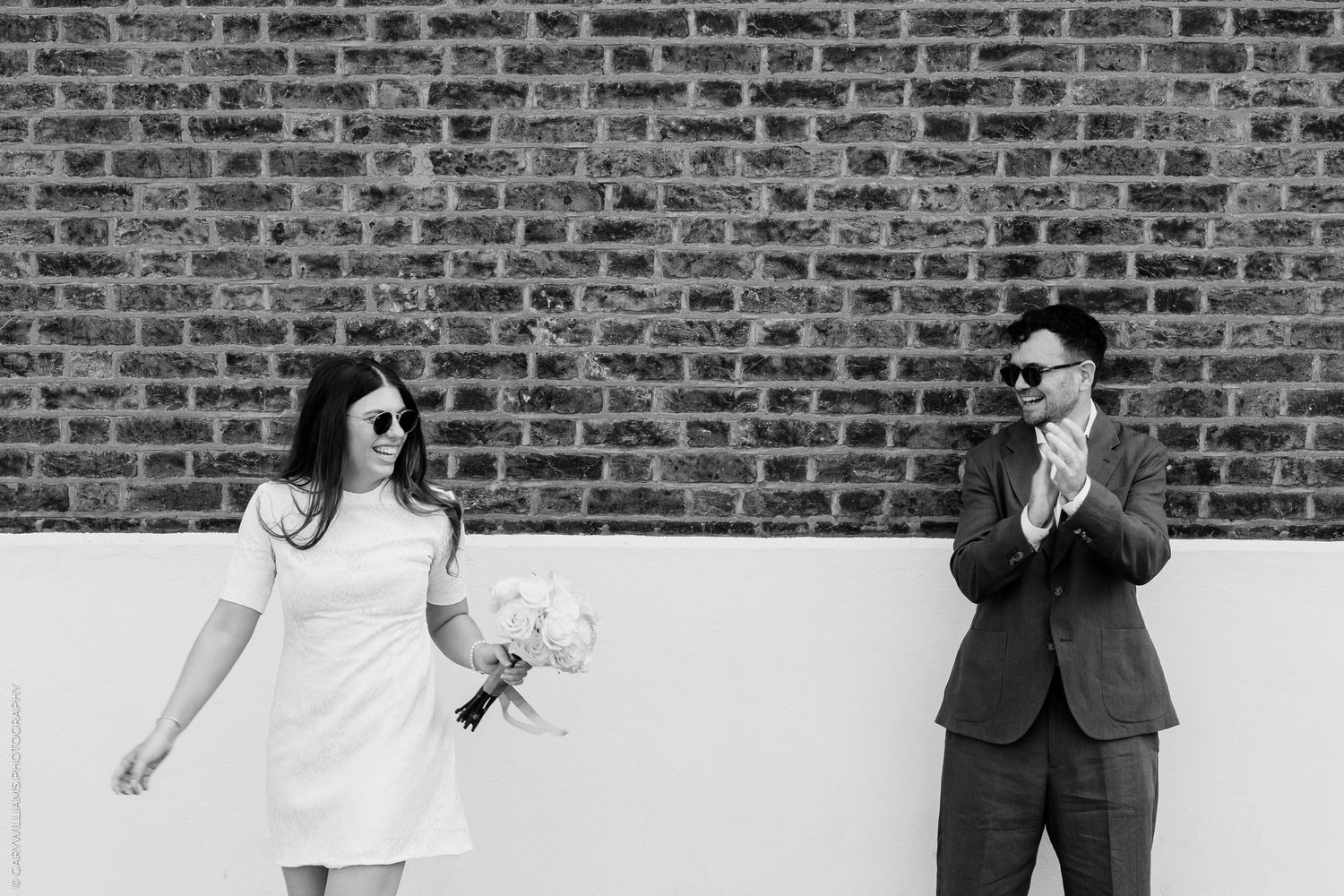 Black and white photo of a bride in short dress holding bouquet as groom claps beside a brick wall.