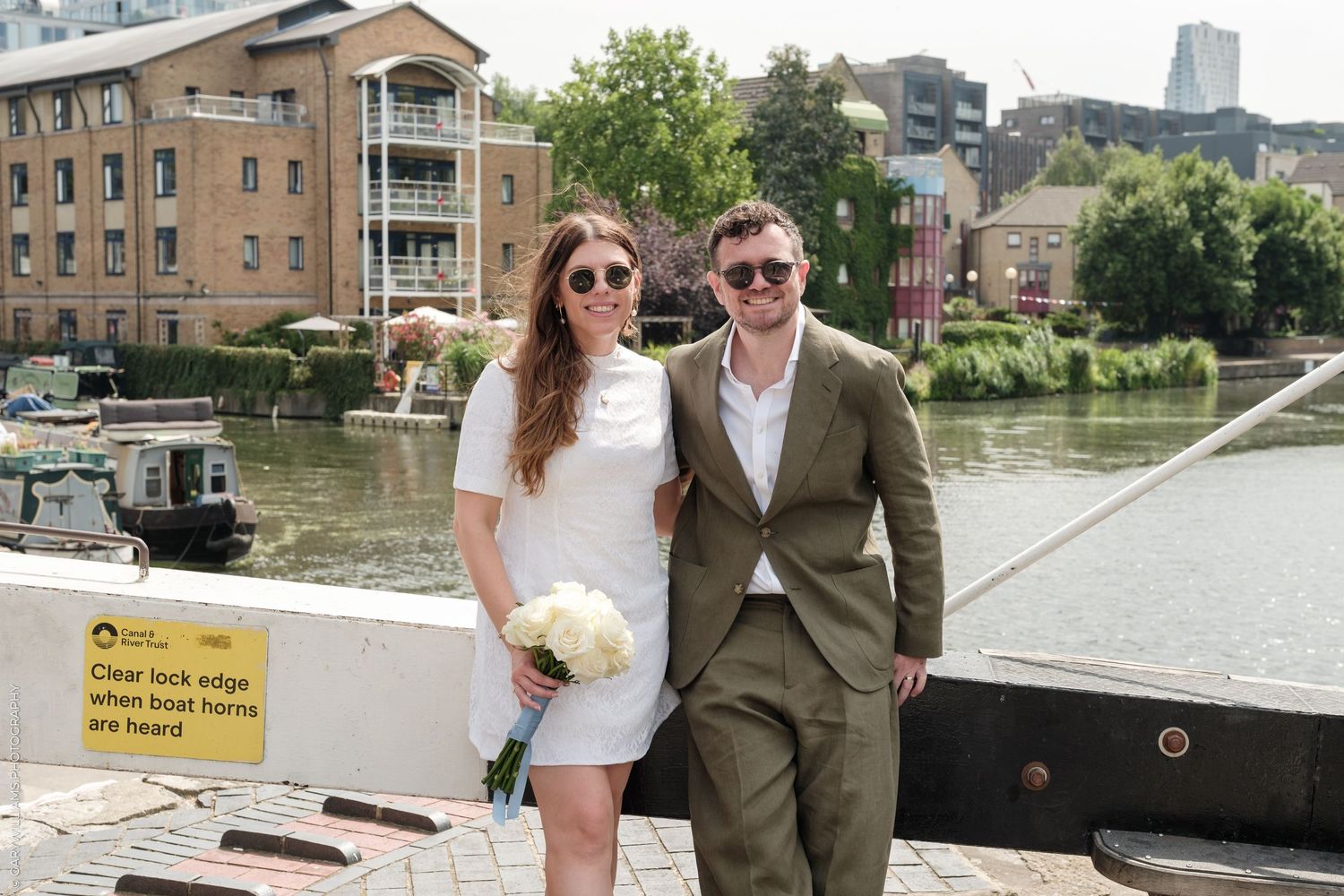 Happy couple posing by a canal lock in London on their wedding day, bride holding white bouquet.