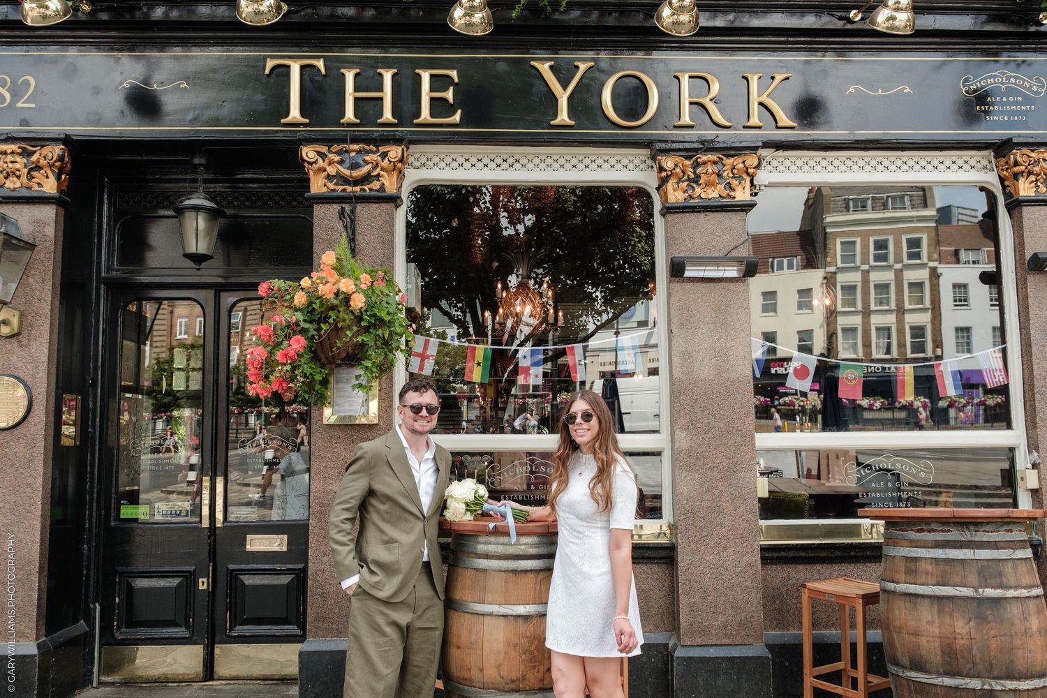 Couple posing outside The York pub in London, woman in white mini dress holding bouquet, man in tan suit.