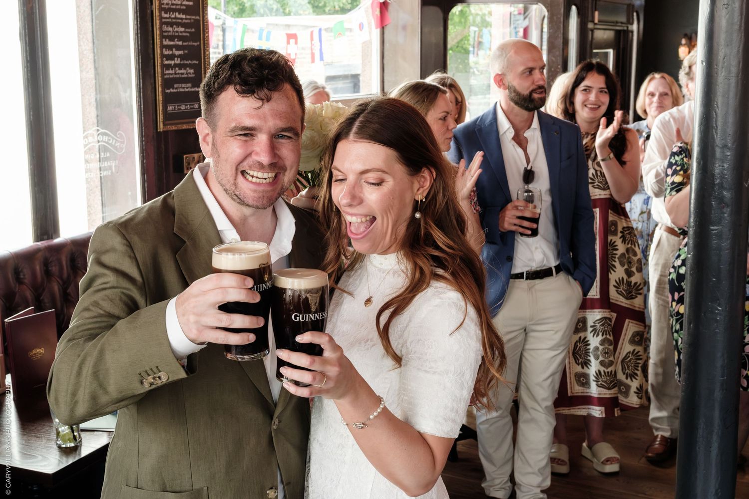 Couple toasting with Guinness pints at a pub, smiling joyfully, with guests celebrating in the background.