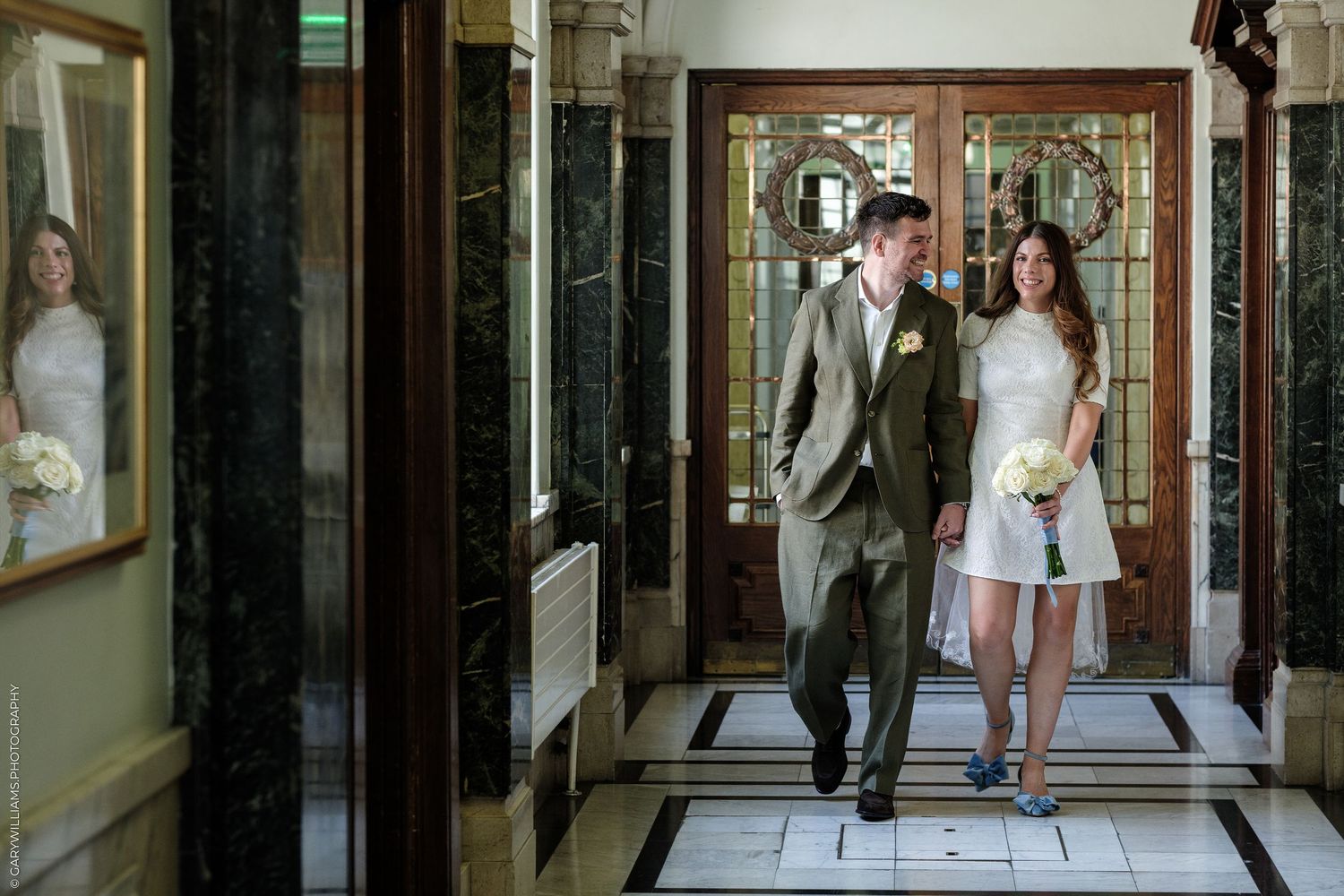 Bride in short white dress and groom in olive suit walking together through elegant marble hallway after wedding ceremony.