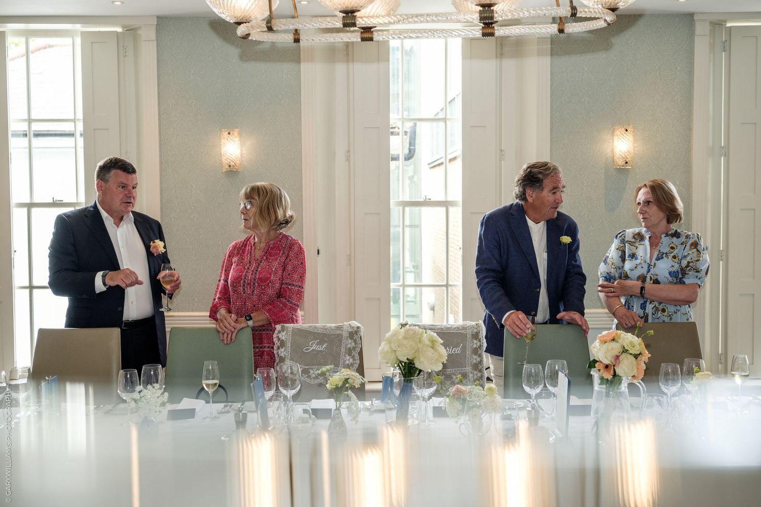 Four elegantly dressed guests socializing at a formal dining table set with glassware in a bright, upscale banquet room.