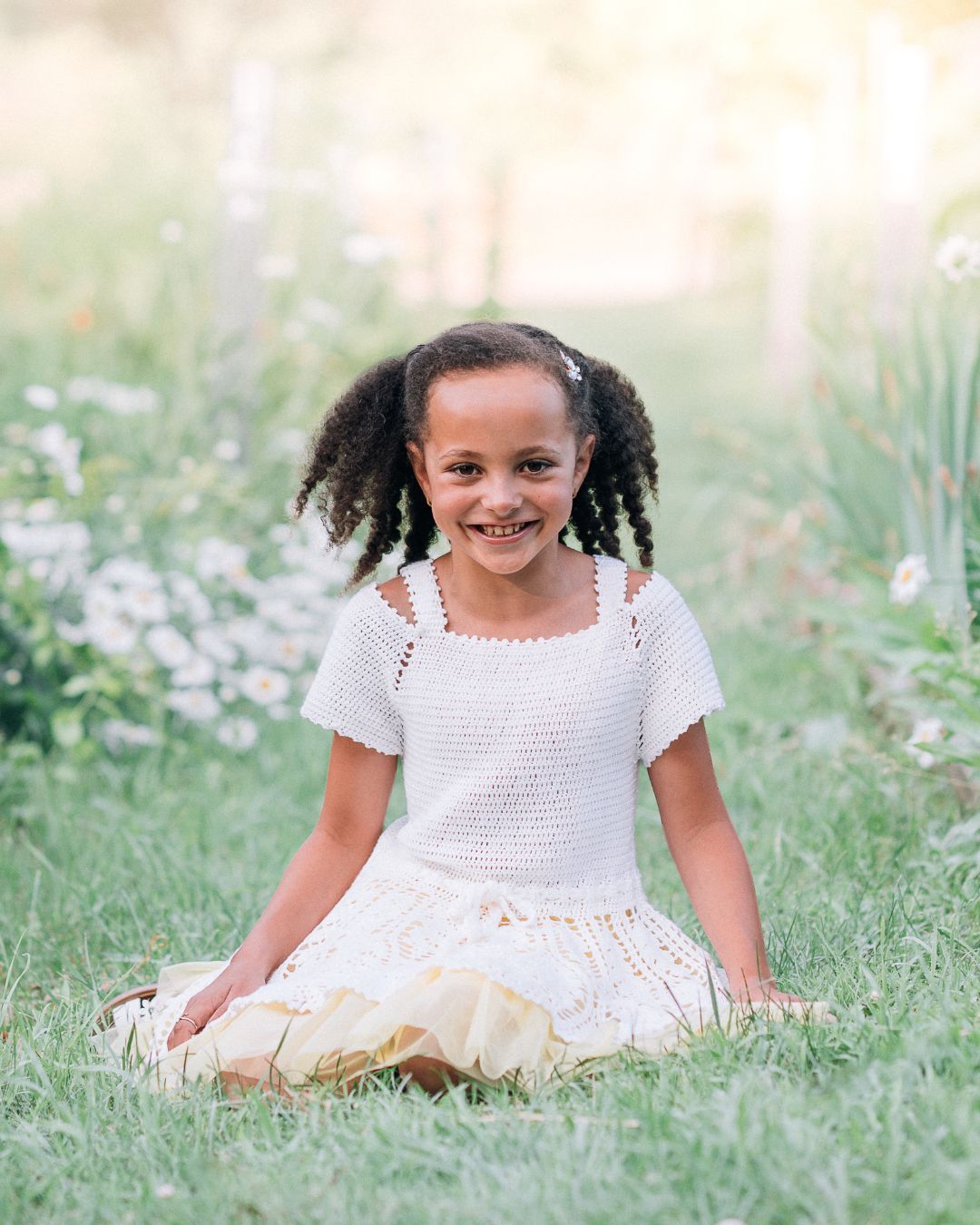 A smiling young girl with curly pigtails sits cross-legged in a lush green meadow wearing a white crochet top and skirt.