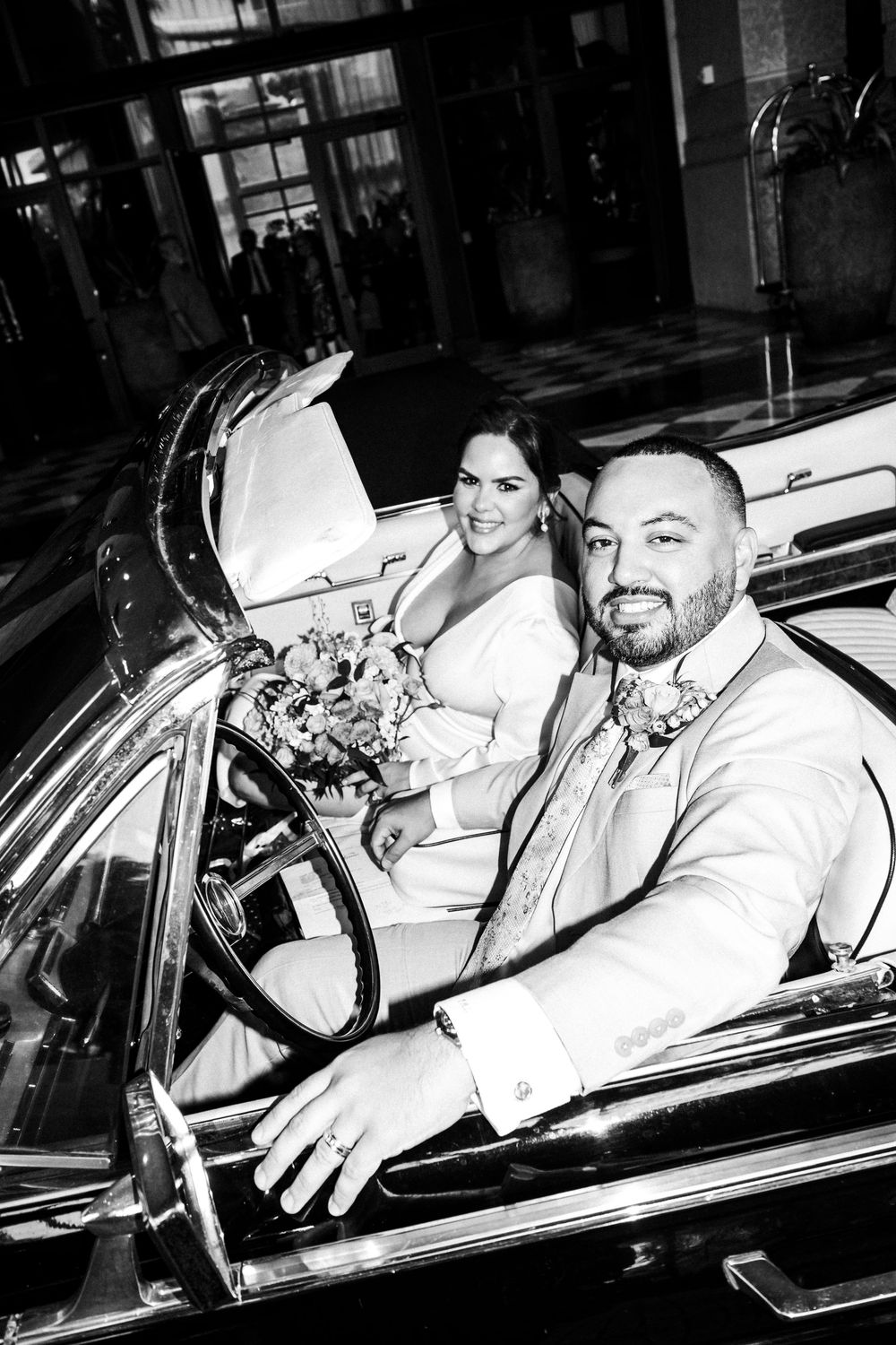Black and white photo of smiling bride and groom in a vintage convertible car on their wedding day.