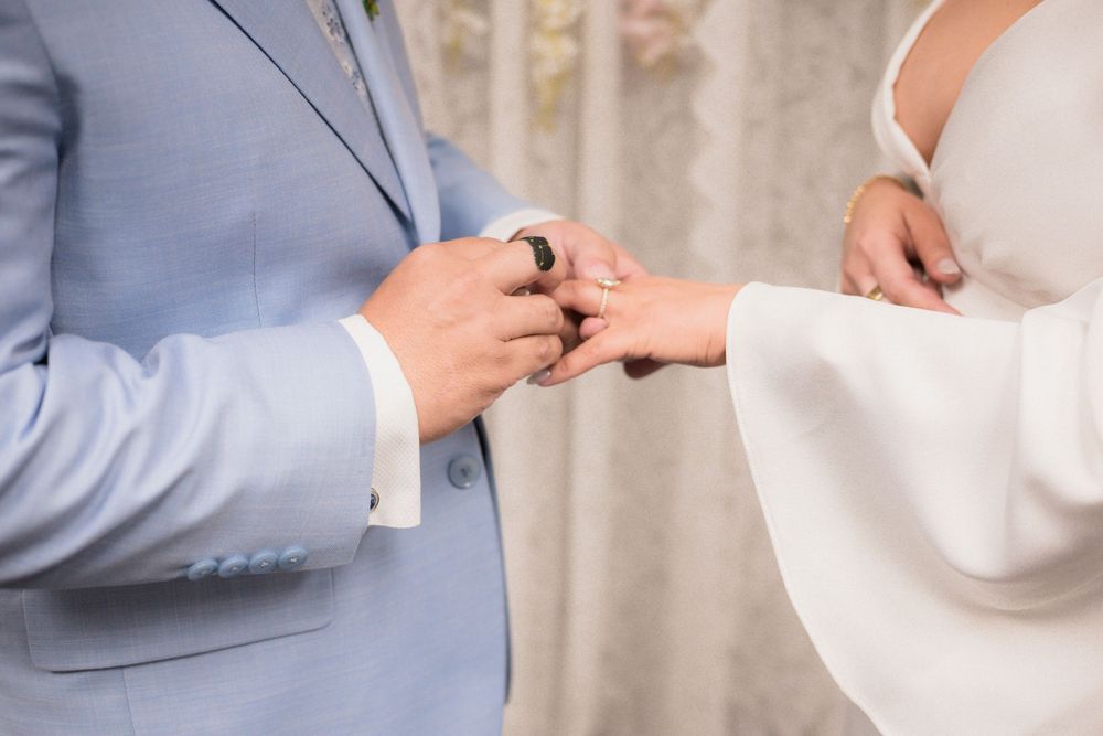 Groom in light blue suit placing a wedding ring on brides finger during a ring exchange ceremony.