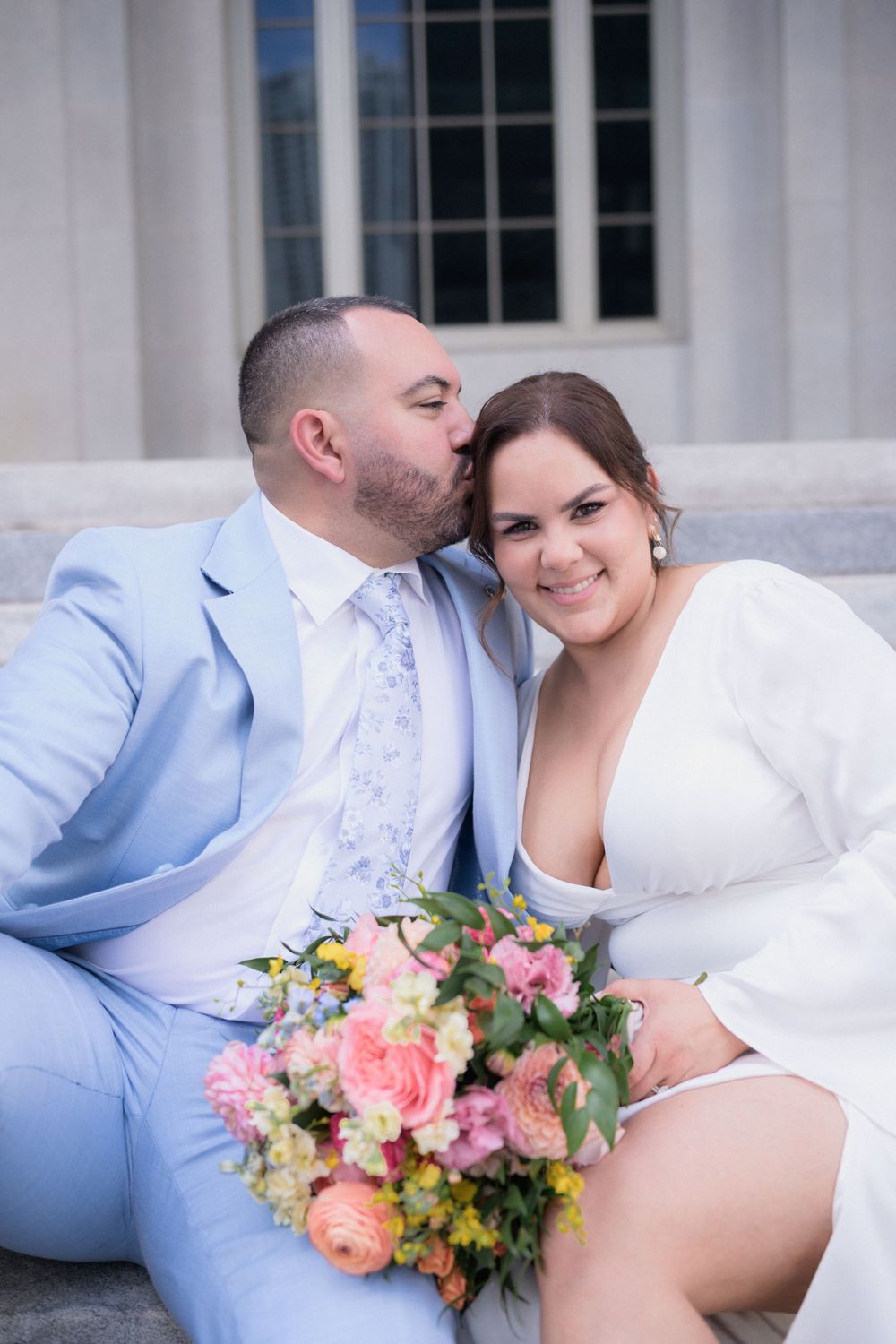 Groom in light blue suit kisses bride in white dress on cheek as she smiles, holding colorful bouquet on steps.