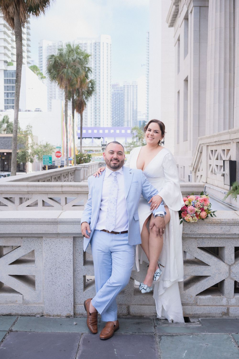 Bride and groom pose outdoors in Miami, she in white dress, he in light blue suit, with palm trees and skyline behind.