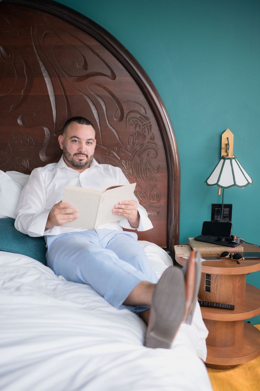 Man in white shirt reading a book while relaxing on a bed with ornate wooden headboard in a teal bedroom.