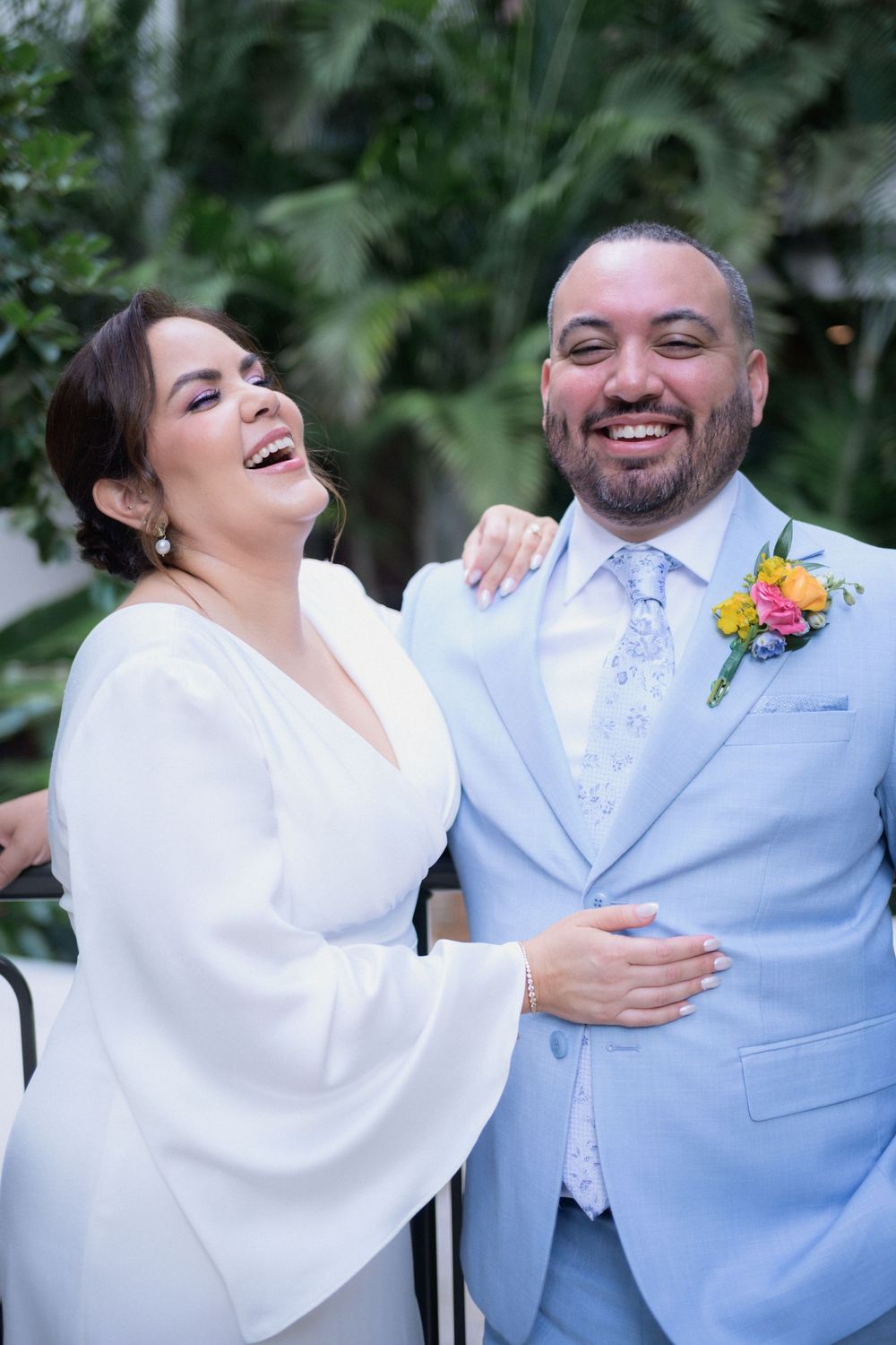 A joyful bride in white and groom in light blue suit with floral boutonniere laughing together at their wedding.