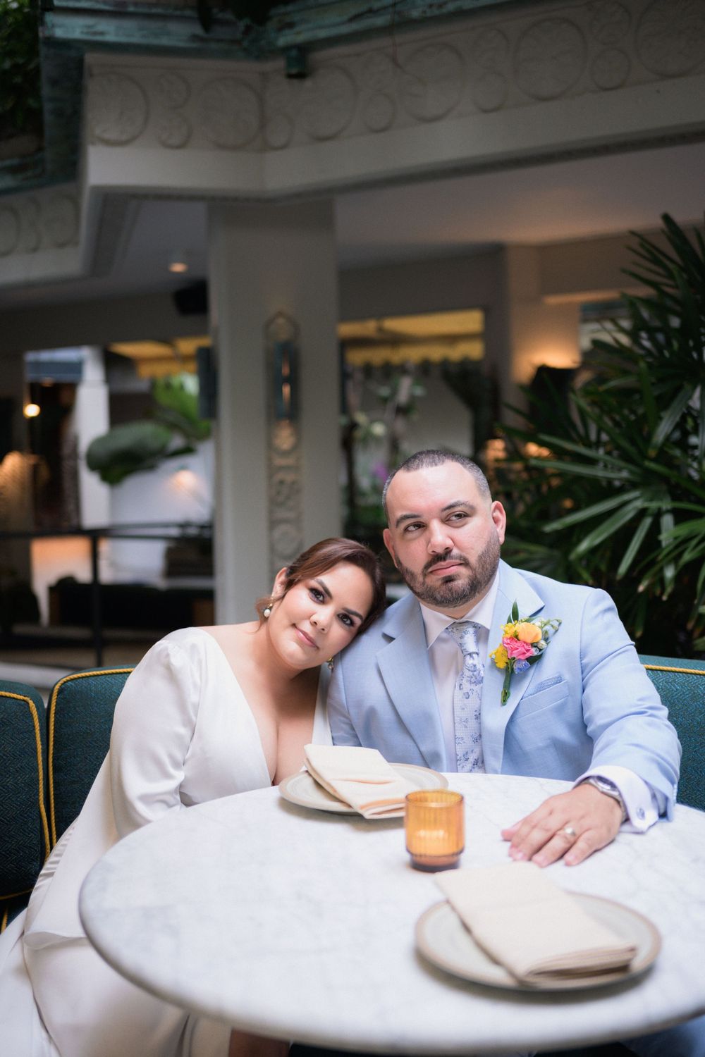 Bride and groom sitting together at a marble table in an elegant restaurant, wearing white dress and light blue suit.