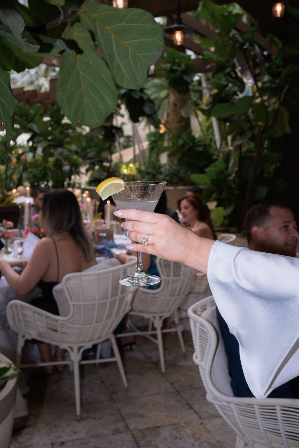 Hand holding a martini glass with lemon garnish at an elegant outdoor restaurant patio surrounded by tropical greenery.