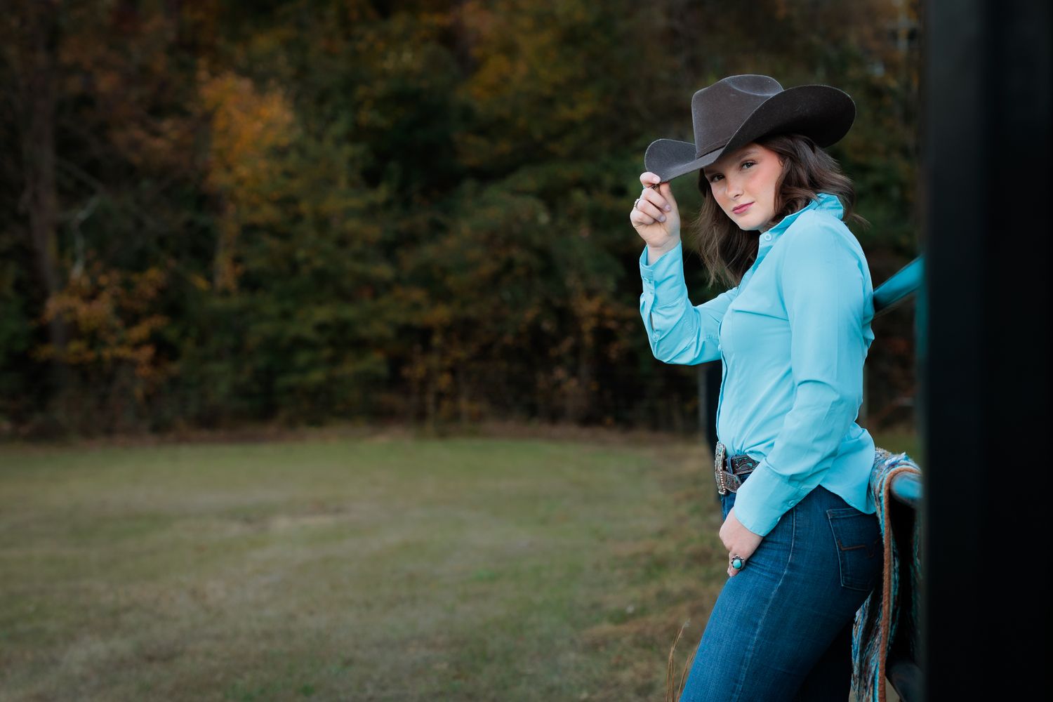 A young woman in a teal shirt and cowboy hat leans against a fence in a rural outdoor setting.