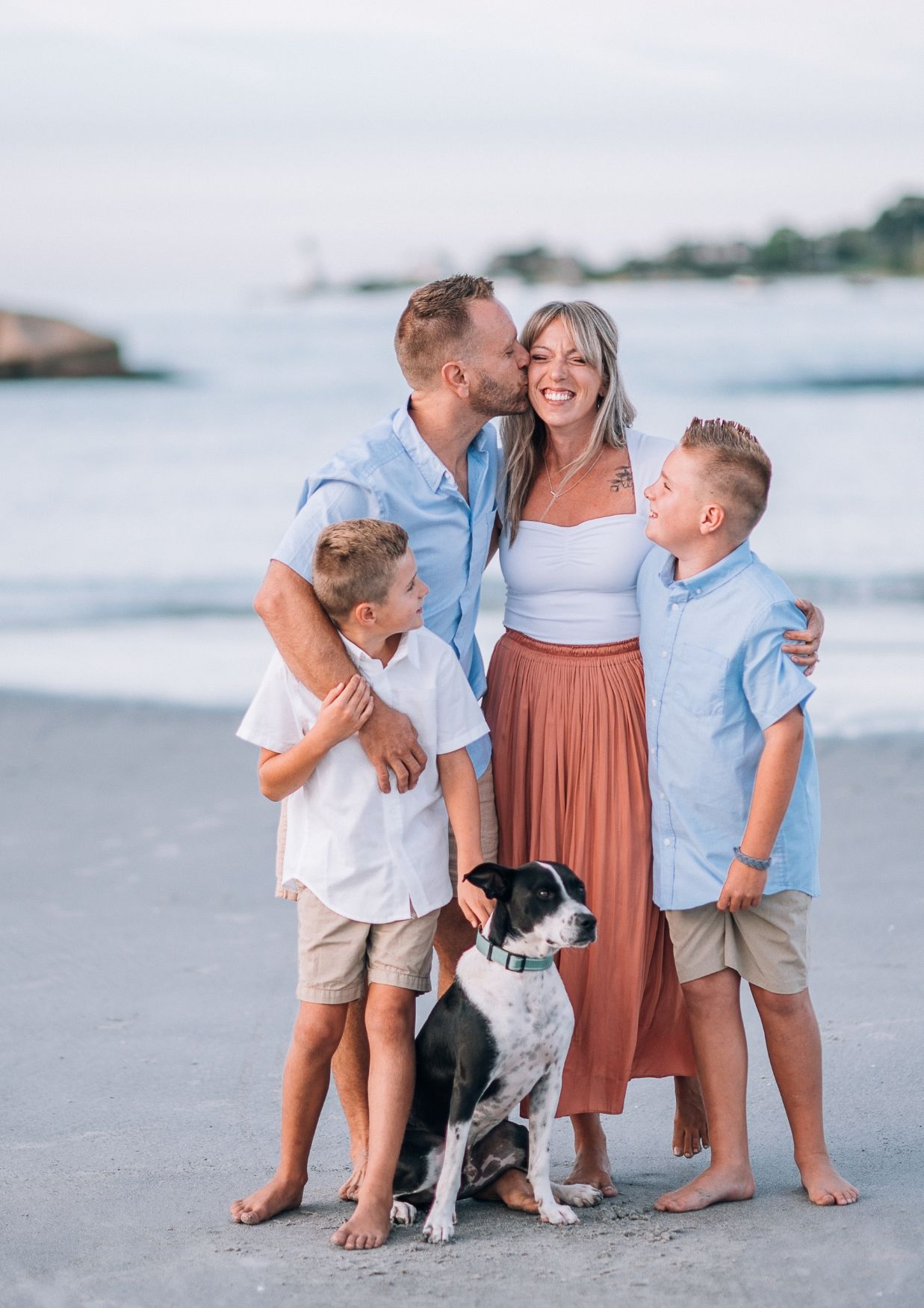 Family of four with dog posing on beach, parents kissing while two boys smile, casual summer outfits.