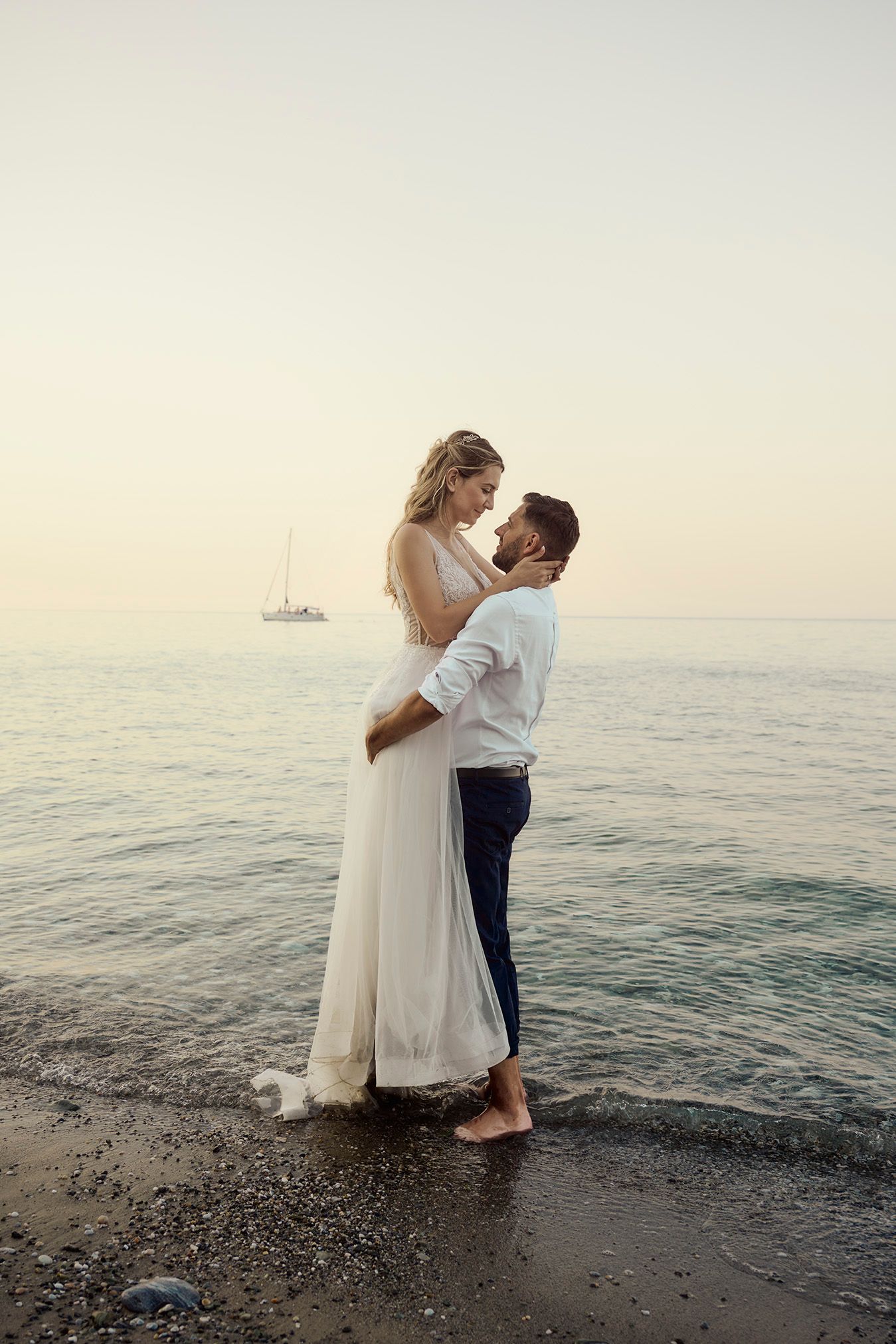 Romantic couple in white embracing on a sandy beach with a sailboat at sunset.