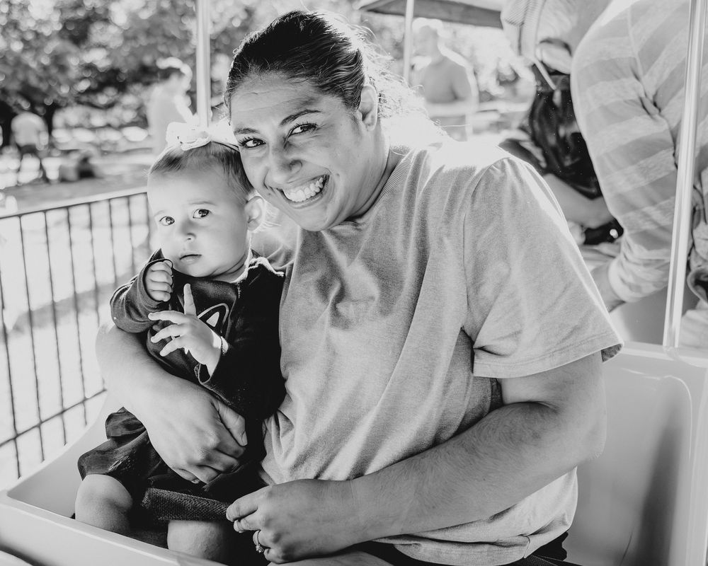 A smiling woman holds a baby on a small ride in a black and white outdoor photo.
