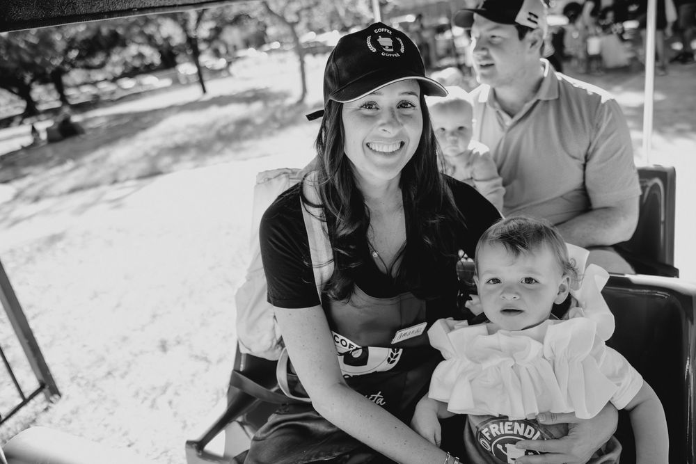 Black and white photo of a smiling woman in a cap holding a baby girl, with a man seated behind them outdoors.