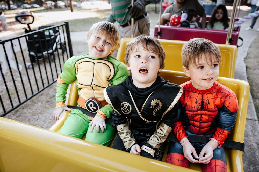 Three young boys in Halloween costumes ride an amusement park ride dressed as Ninja Turtle, Power Ranger, and Spider-Man.