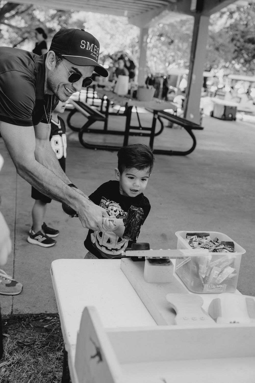 Man in baseball cap helps young child at outdoor picnic table during Halloween party.