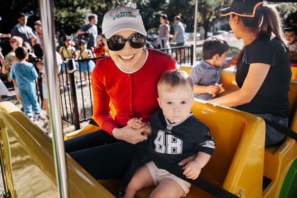 Woman in red cardigan and sunglasses holds toddler in football jersey on amusement park ride outdoors.