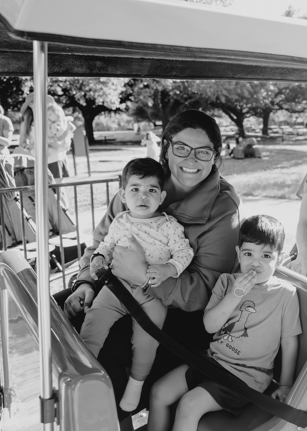 Black and white photo of a woman with glasses holding a baby and sitting with a young boy in an outdoor vehicle.