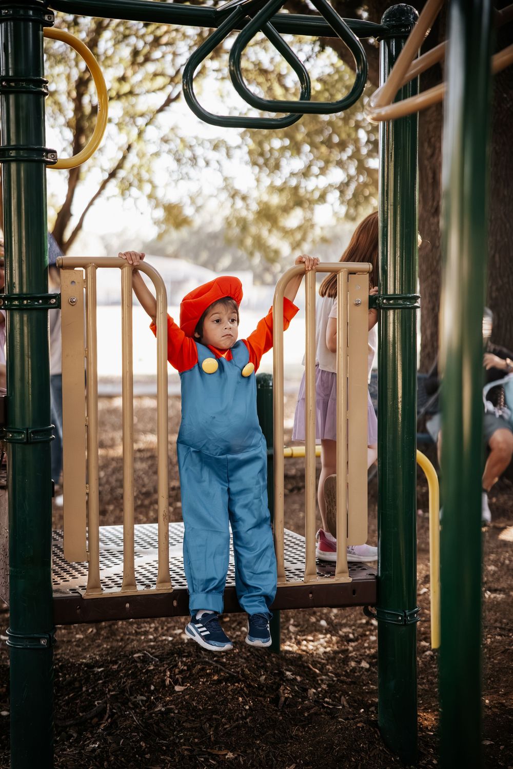 A young child in a Mario costume plays on a colorful playground structure outdoors.