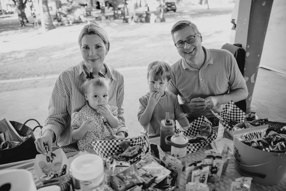 A smiling family with two young children sits together at a table with food and crafts.