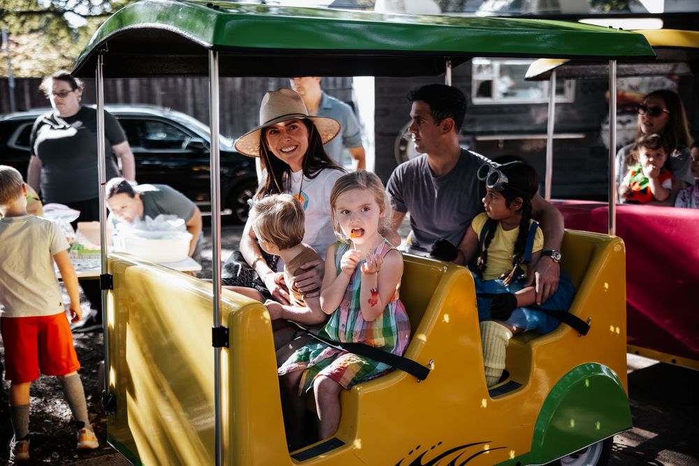 Families with young children riding a colorful yellow train ride outdoors on a sunny day at a festive event.