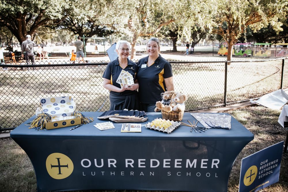 Two staff members smile at an Our Redeemer Lutheran School information booth outdoors at a community event.