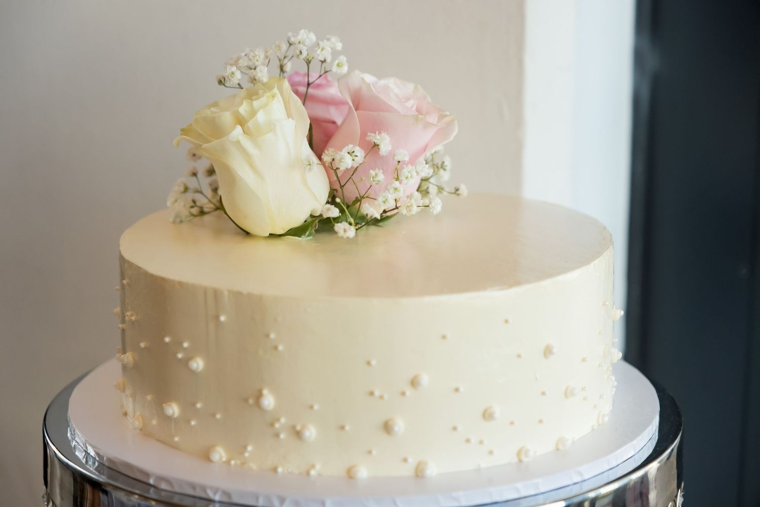 Elegant single-tier white wedding cake with pearl details, topped with fresh pink and cream roses and baby's breath.