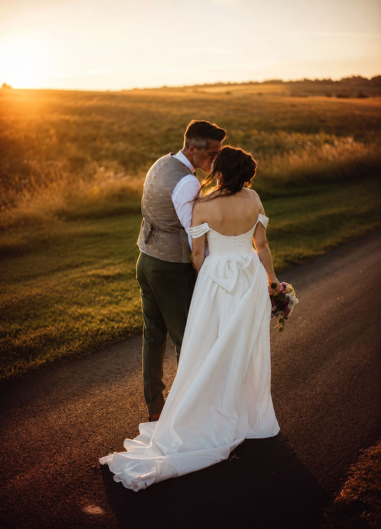 Bride and groom sharing a kiss at golden hour sunset on a country road, bride in elegant white gown with bow.