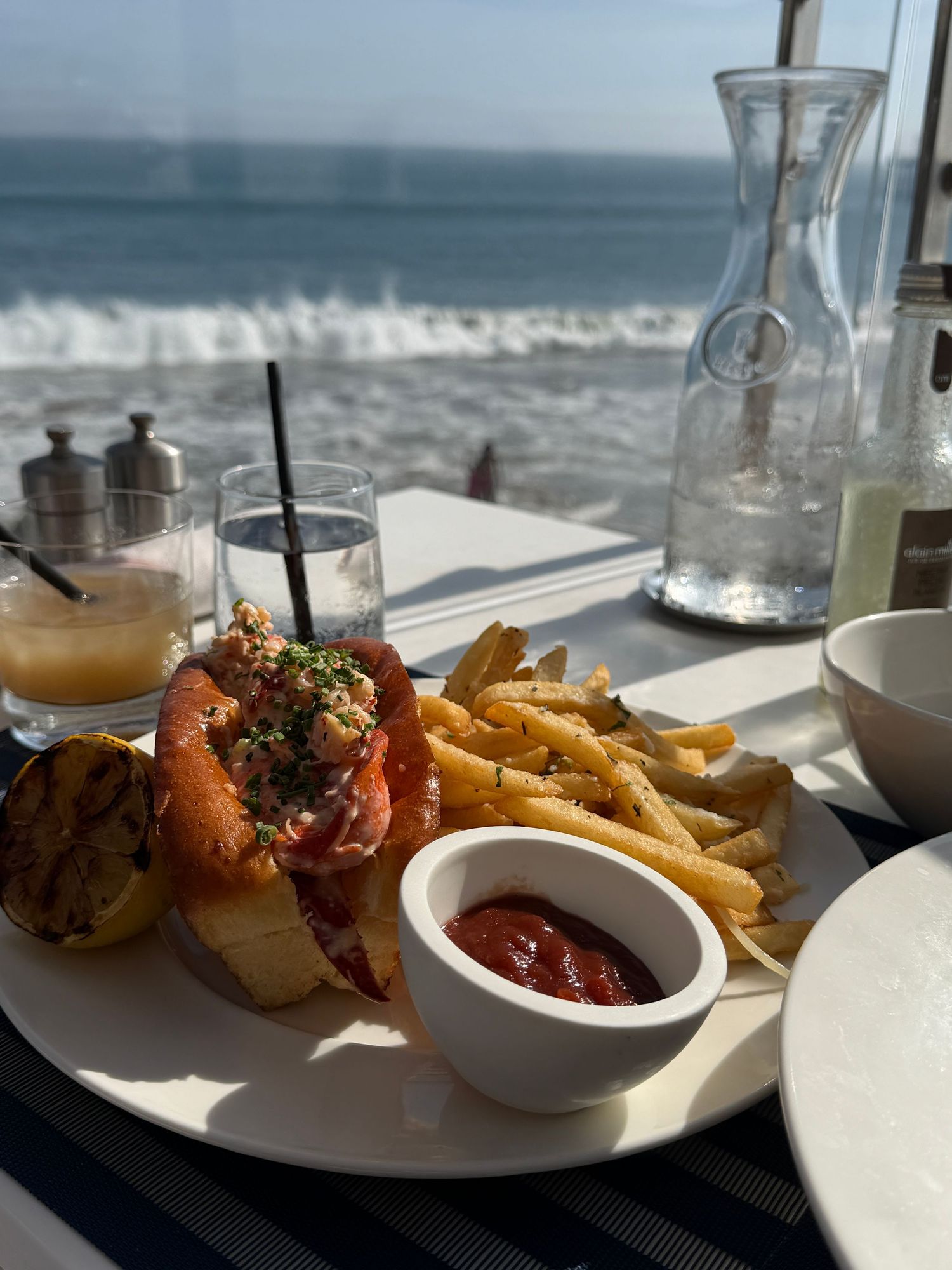 Lobster roll with golden fries and ketchup served on a white plate with ocean views at a beachside restaurant.